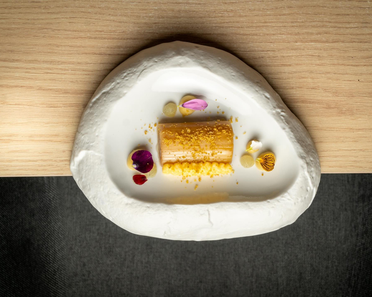 Puerro confitado a baja temperatura, acompau00f1ado de una emulsiu00f3n de pepinillo y aceituna, terminado con una holandesa delicadamente emulsionada y una tierra fina de aceituna.
